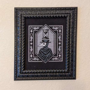 May include: Black and white cross-stitch embroidery in a black frame. The embroidery depicts a plague doctor wearing a long black coat and a mask with a beak. An hourglass sits on top of the doctor's head.