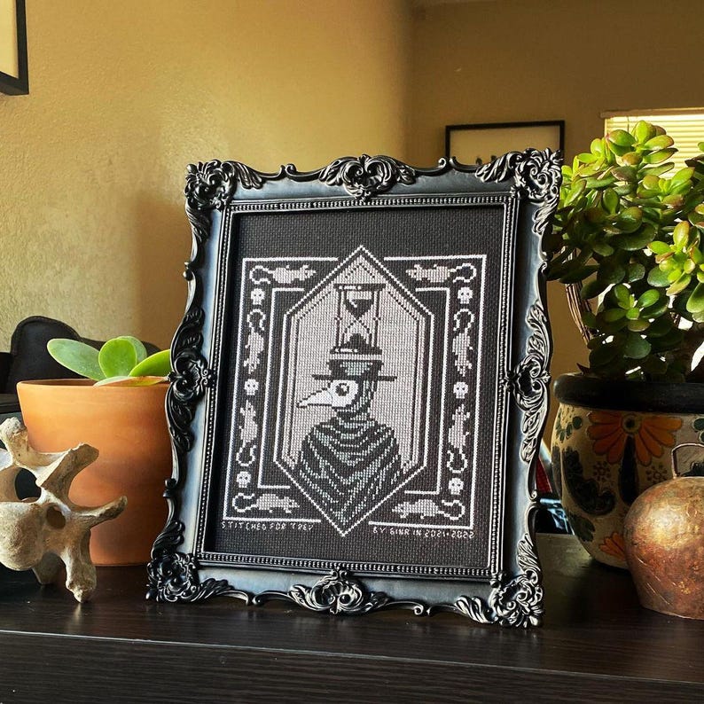 May include: Black and white cross-stitch embroidery in a black frame depicting a plague doctor wearing a mask and hat with an hourglass on top. The embroidery is titled "Stitched for Thy" and "KY Birk in 2001-2002".