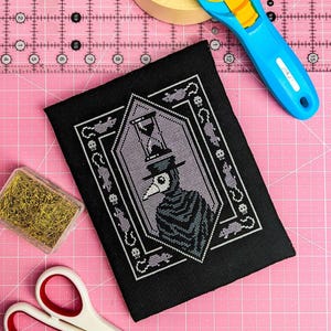 May include: Black cross-stitch embroidery on black fabric with a white and purple design of a figure wearing a plague doctor mask and hat. The figure is holding an hourglass. The design is surrounded by a black frame with white and purple details.