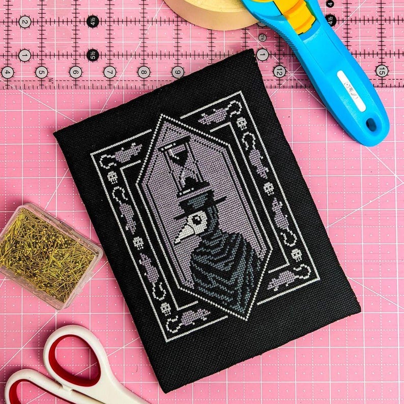 May include: Black cross-stitch embroidery on black fabric with a white and purple design of a figure wearing a plague doctor mask and hat. The figure is holding an hourglass. The design is surrounded by a black frame with white and purple details.