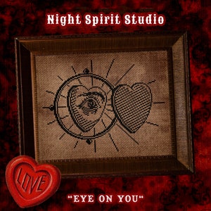 May include: A framed cross-stitch design featuring a heart with an eye inside and a second heart. The design is on a brown background and the frame is brown wood. The text "EYE ON YOU" is below the frame. A red heart with the word "LOVE" embossed on it is in the bottom left corner.