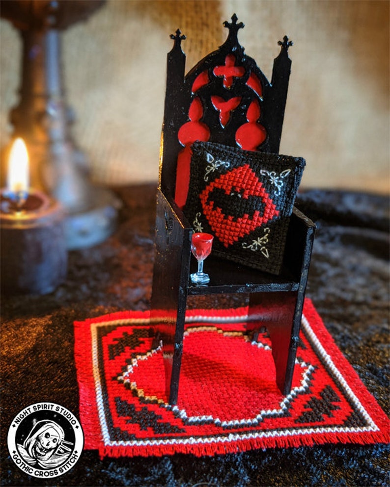 May include: A miniature black gothic chair with a red cushion and a bat-shaped pillow. A small glass of red liquid sits on a shelf on the chair. The chair is on a red and black rug with a white border. The rug has a fringe border. The image includes the text "Night Spirit Studio Gothic Cross Stitch".