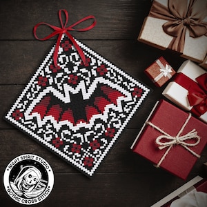 May include: A black and red cross stitch pattern of a bat with a white border. The pattern is on a square piece of fabric and is hanging from a red ribbon. The fabric is on a dark wood surface. The image also includes a logo for "Night Spirit Studio Gothic Cross Stitch".