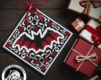 May include: A black and red cross stitch pattern of a bat with a white border. The pattern is on a square piece of fabric and is hanging from a red ribbon. The fabric is on a dark wood surface. The image also includes a logo for "Night Spirit Studio Gothic Cross Stitch".