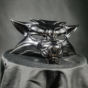 Peut inclure: Un masque de loup argenté métallique avec des oreilles pointues, un museau et des crocs visibles. Le masque est présenté sur une surface noire sur un fond gris foncé. La surface du masque reflète la lumière, soulignant ses détails sculptés.