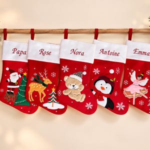 Op de afbeelding: Vijf rode kerstsokken met witte boorden, elk geborduurd met de namen Papa, Rose, Nora, Antoine en Emma. De sokken zijn voorzien van applicaties van de kerstman, een rendier, een teddybeer, een pinguïn en een fee, met sneeuwvlokken.