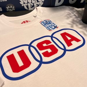 以下が含まれることがあります： 白地に青と赤の「USA」のグラフィックがプリントされたTシャツ。文字は青で縁取りされ、赤で塗りつぶされています。