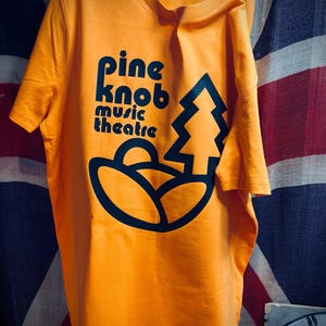 Op de afbeelding: Een oranje t-shirt met korte mouwen en een donkerblauwe grafische print. Het ontwerp bevat de tekst "pine knob music theatre" in een afgerond lettertype, vergezeld van een gestileerde dennenboom en een gebogen landschapsvorm onder de tekst.