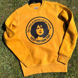 Puede incluir: Una sudadera de color amarillo mostaza con un gráfico circular negro. El gráfico presenta a una persona con el pelo rizado y el texto "The litter problem is in our hands. Keep Britain Tidy."