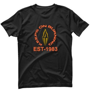 Peut inclure: T-shirt noir avec un graphique circulaire orange qui dit "Keeps On Burning" avec un motif de flamme au centre et "EST-1983" en dessous.