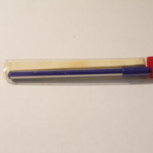 May include: A blue and white striped pen with a red cap is inside a clear plastic tube. A rectangular, off-white eraser is also inside the tube. The pen and eraser are on a white surface.