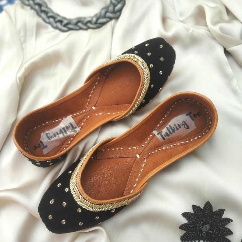 May include: A pair of black velvet shoes with gold trim and black sequins. The shoes have a pointed toe and a low heel. The shoes are labeled "Talking Toe".