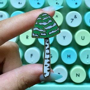 May include: A green and white enamel mushroom pin with a gold outline. The mushroom has a white stem with black markings that resemble a birch tree.