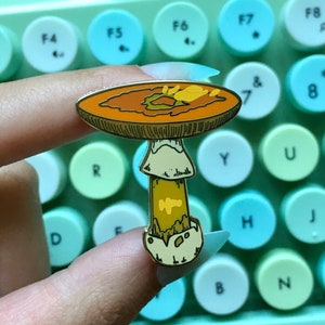 May include: A gold-tone mushroom enamel pin with a brown cap and yellow stem. The pin has a white base and a detailed design.