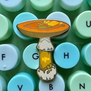 May include: A gold-tone enamel pin of a mushroom with an orange cap and a yellow stem. The cap has a brown and yellow pattern. The pin is on a green and blue background.