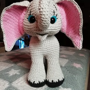 Puede incluir: Un juguete de elefante de crochet gris con orejas rosas y ojos azules. El elefante está de pie sobre una manta gris y blanca.