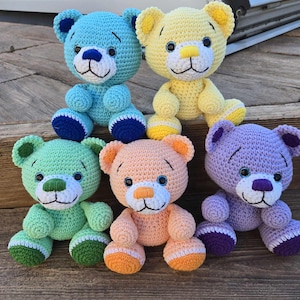 May include: Five crocheted teddy bears in different colours: light blue, yellow, green, peach, and purple. Each bear has embroidered eyes, a nose, and a mouth.