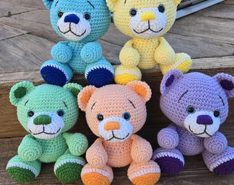 May include: Five crocheted teddy bears in different colors: light blue, yellow, green, peach, and purple. Each bear has embroidered eyes, a nose, and a mouth.