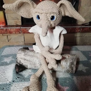 May include: A crocheted doll with large ears, blue eyes, and a long nose, wearing a white cloth. The doll is sitting on a log. The text "Dobrá klubka z Dobré" is at the top of the image.