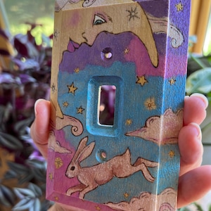 May include: A wooden light switch cover with a whimsical design featuring a crescent moon with a face, stars, clouds, and a rabbit hopping in the sky. The background is painted in shades of purple, blue, and pink.