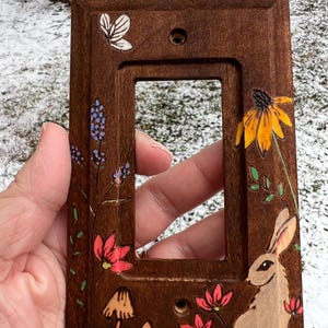 May include: A wooden light switch cover with hand-painted floral and woodland designs. The cover features a rabbit, flowers, mushrooms, and a butterfly. The color palette includes brown, red, yellow, blue, and green accents.