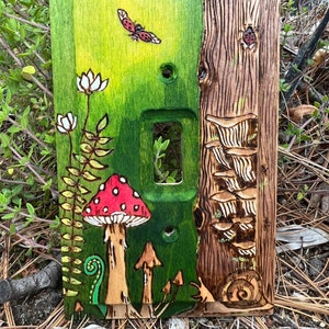 May include: A wooden light switch cover with a hand-painted forest scene. The cover features a red and white mushroom, green ferns, a ladybug, and a tree trunk with mushrooms growing on it.