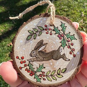 May include: A round wooden ornament with a painted brown rabbit leaping across the center. The ornament is decorated with green holly leaves and red berries around the edges. A natural twine loop is attached for hanging.