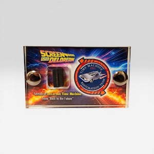 May include: A clear acrylic display case featuring a section of the DeLorean Time Machine from "Back to the Future." The case has a colorful background with the text "SCREEN USED DELOREAN" and "TIME MACHINE 1985." Two silver spheres are on the front.