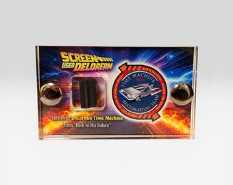 Puede incluir: Una vitrina de acrílico transparente con una sección de la máquina del tiempo DeLorean de "Regreso al futuro". La vitrina tiene un fondo colorido con el texto "SCREEN USED DELOREAN" y "TIME MACHINE 1985". Dos esferas plateadas están en el frente.