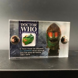 May include: A clear plastic display case featuring a green and brown Ice Warrior head from the Doctor Who television series. The case includes a small piece of the original prop used in the show. The text on the case reads "DOCTOR WHO" and "A piece from an original Ice Warrior head seen in Doctor Who between 1967-1974."
