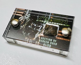 May include: A clear acrylic display case with a section of screen used in the movie "Alien Resurrection". The case has two silver metal knobs on the sides and a black and green image of the alien creature from the movie. The text "Alien Resurrection" is printed on the case.