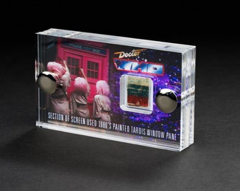 May include: A clear acrylic display case with a section of screen used in the 1980's painted TARDIS window pane. The case has a blue and purple background with the words "Doctor Who" printed on it. The case also has two silver metal knobs on the sides.