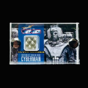 May include: A clear plastic display case with a silver section of a screen-used Cyberman from the Doctor Who television series. The case has a blue background with the text "Doctor Who" and "Silver Nemesis" printed on it. The case also has two silver metal fasteners on the sides.