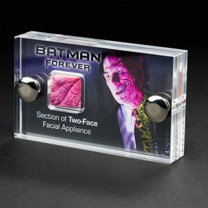 May include: A clear acrylic display case containing a section of Two-Face's facial appliance from the film Batman Forever. The case also features a photograph of Two-Face, with the text "Batman Forever" above the image.
