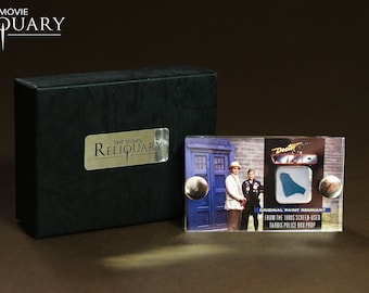May include: A clear plastic card with a blue paint chip, a photo of the TARDIS, and two actors from the Doctor Who television series. The card has the text "The Movie Reliquar", "Doctor Who", "Original Paint Remnant", "From the 1980s Screen-Used TARDIS Police Box Prop".