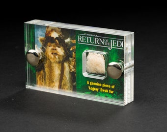 Könnte beinhalten: Eine transparente Acryl-Vitrine mit einem Star Wars: Rückkehr der Jedi-Ritter-Bild von Chewbacca. Die Vitrine enthält ein kleines Quadrat aus hellfarbenem Fell und den Text: "Ein echtes Stück 'Logray' Ewok-Fell."