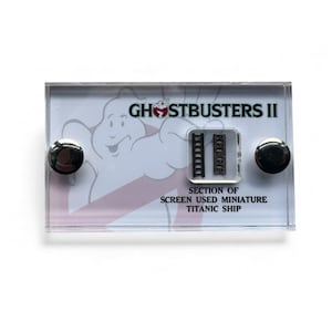 Peut inclure: Un présentoir en acrylique transparent avec le logo Ghostbusters II et un fantôme de dessin animé. Il comprend deux petites échelles miniatures noires et le texte "SECTION OF SCREEN USED MINIATURE TITANIC SHIP". Deux aimants ronds argentés fixent le présentoir.