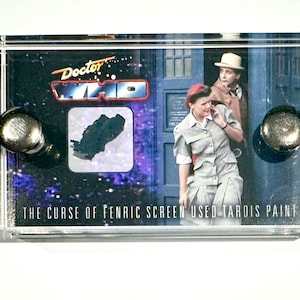 May include: A clear acrylic display case with a black and white image of a scene from the Doctor Who television series. The image shows two characters, a woman and a man, standing in front of a blue police box. The text "Doctor Who" is printed in red and white on the top of the case. The text "The Curse of Fenric Screen Used Tardis Paint" is printed on the bottom of the case.