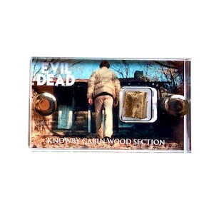 Op de afbeelding: Een heldere acryl display case met een goudkleurig houten gedeelte. De case bevat een afbeelding van een persoon die naar een hut loopt, met de tekst "EVIL DEAD" en "KNOWBY CABIN WOOD SECTION".