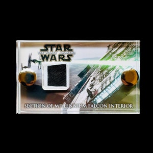 May include: A clear plastic display case with a black fabric sample inside. The case has a gold background with the text "STAR WARS THE FORCE AWAKENS" and "SECTION OF MILLENNIUM FALCON INTERIOR".