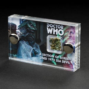 Könnte beinhalten: Eine transparente Acryl-Vitrine mit einem Teil des Bildschirms, der in der Doctor Who-Episode "Sea Devil" aus dem Jahr 1972 verwendet wurde. Die Vitrine zeigt ein grünes und braunes Bildschirmstück, ein Schwarzweißfoto einer Kreatur und den Text "Doctor Who".