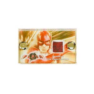 Könnte beinhalten: Nahaufnahme eines roten und gelben Stoffmusters vom Flash-Kostüm. Das Muster ist in einem durchsichtigen Plastikrahmen mit goldenem Hintergrund ausgestellt. Der Text "Section of The Flash Costume" ist unter dem Muster gedruckt.