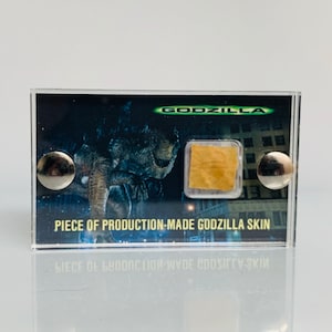 Può includere: Una teca di plastica trasparente che contiene un pezzo di materiale marrone etichettato "PIECE OF PRODUCTION-MADE GODZILLA SKIN" con un'immagine in bianco e nero di Godzilla sullo sfondo. La teca ha un bottone argentato su ogni lato.