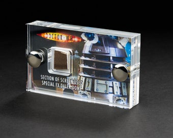 May include: A clear acrylic display case with a small piece of brown material inside. The case has a silver background with a picture of a Dalek from the Doctor Who television series. The text on the case reads "Doctor Who" and "Section of Screen Used Special FX Dalek Dome".