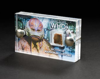 May include: A clear plastic display case with a brown piece of fabric labeled "Section of Marshman Costume" and a photo of a creature from the Doctor Who TV series. The text "Doctor Who" is printed on the case.