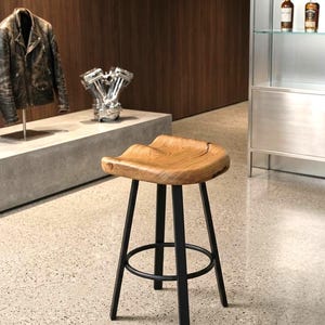 Può includere: Uno sgabello da bar con seduta sagomata in legno chiaro naturale con venature visibili e una crepa sul lato destro. Lo sgabello è sostenuto da quattro gambe dritte in metallo nero collegate da un poggiapiedi circolare in metallo nero.