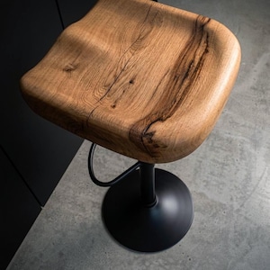 Handcrafted solid wood barstool with a contoured saddle seat showing natural wood cracks. Black metal loft-style frame and base. Two minimalist counter height stools in a warm neutral kitchen setting with industrial design elements.