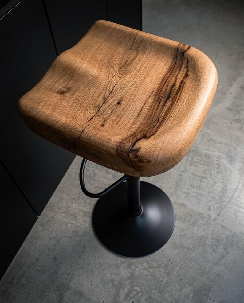 Handcrafted solid wood barstool with a contoured saddle seat showing natural wood cracks. Black metal loft-style frame and base. Two minimalist counter height stools in a warm neutral kitchen setting with industrial design elements.