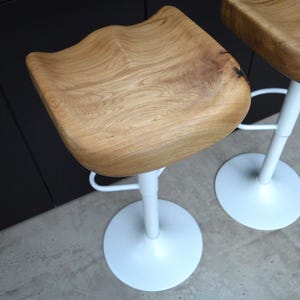 Urban industrial counter stool crafted with a beautifully contoured solid oak saddle seat and a sleek black pedestal stand. Set of two artisanal kitchen counter stools blending rustic woodwork with contemporary design, set against a dark kitchen island with concrete floors.