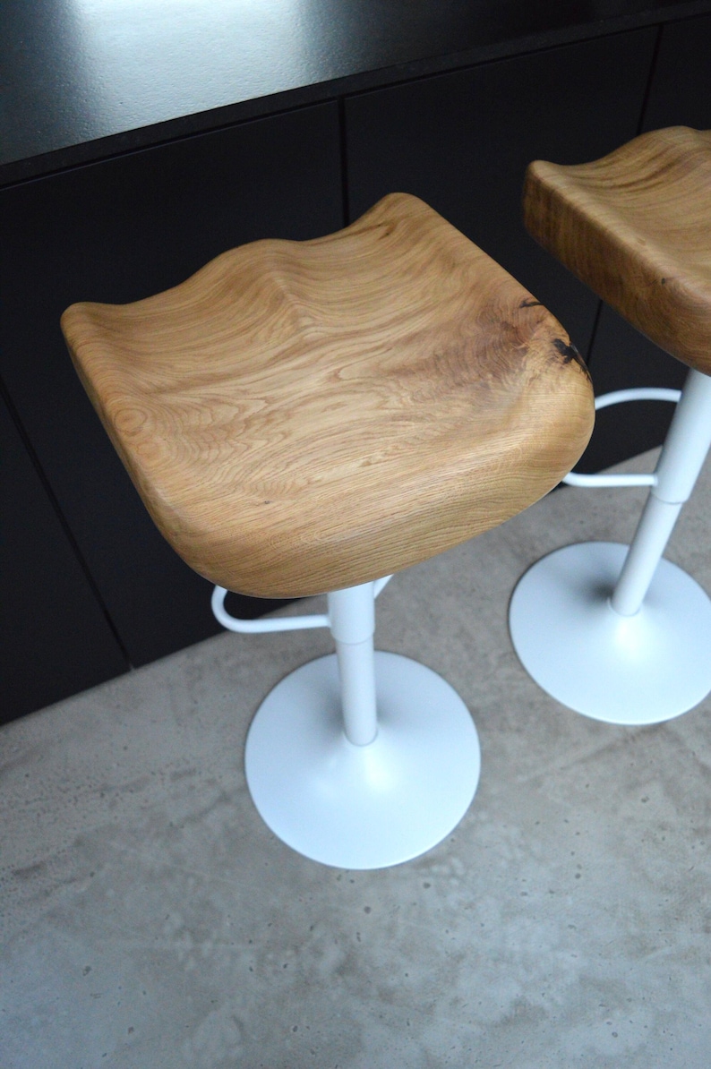 Urban industrial counter stool crafted with a beautifully contoured solid oak saddle seat and a sleek black pedestal stand. Set of two artisanal kitchen counter stools blending rustic woodwork with contemporary design, set against a dark kitchen island with concrete floors.